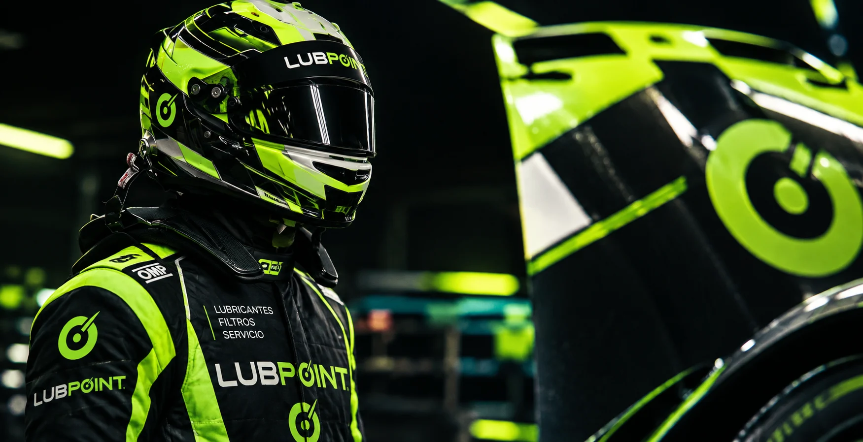 Piloto con traje y casco LUBPOINT junto a un auto con la gráfica verde de LUBPOINT, en un box oscuro.